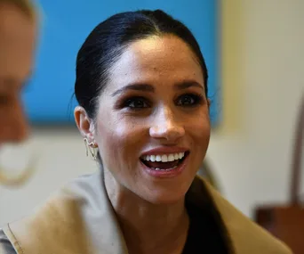 Meghan Markle smiling, wearing earrings and a light-colored coat, with a blue background behind her.