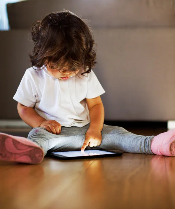 A toddler sitting on the floor uses a tablet, fully focused on the screen.