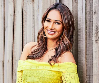 Smiling woman with long hair in a yellow lace dress standing against a wooden fence.