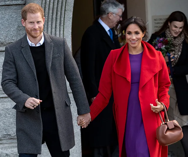 Couple holding hands, smiling, outside a building; woman in red coat and purple dress, man in grey coat.