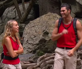 Pair in red tank tops and beige pants talking outdoors beside large rocks and roots, appearing cheerful.