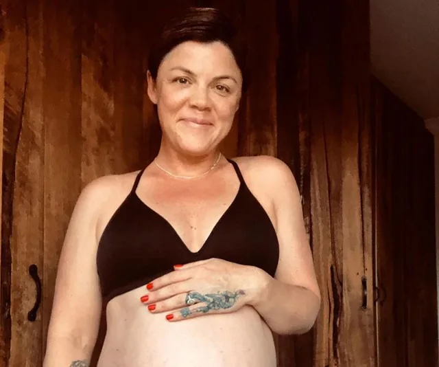 A person in a black top, with short hair, smiles while holding their pregnant belly. Wood panel background.
