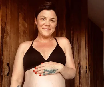 A person in a black top, with short hair, smiles while holding their pregnant belly. Wood panel background.