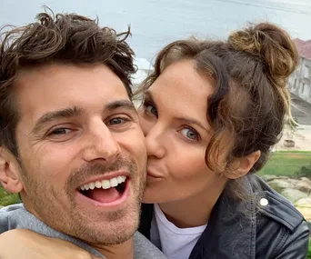 A couple smiling and taking a selfie outdoors with a scenic background.