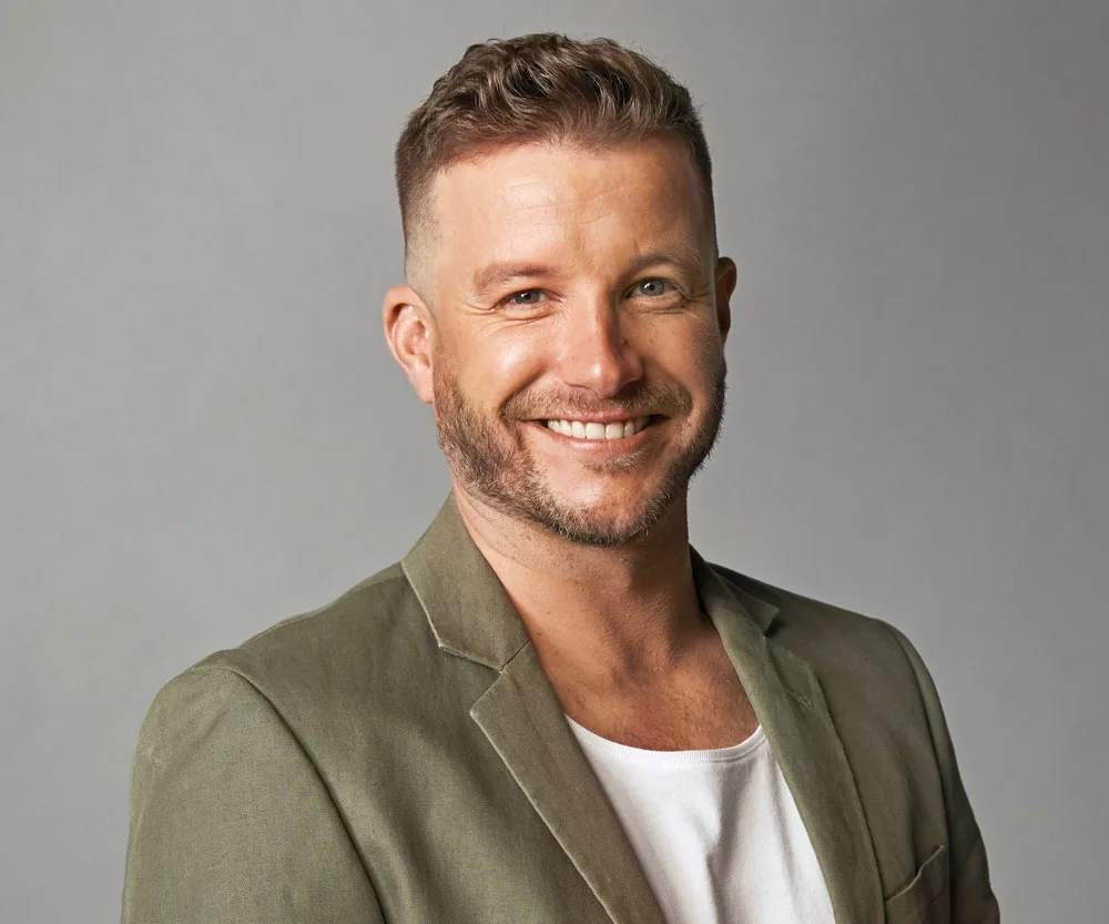 A person smiling, wearing a green blazer and white shirt against a gray background.