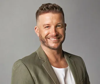 A person smiling, wearing a green blazer and white shirt against a gray background.