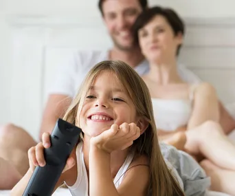 A smiling child with a remote control, lying in front of two blurred adults on a bed, enjoying family TV time.