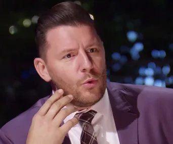 Man in a suit with an animated expression, gesturing with his hand, in a dimly lit setting.