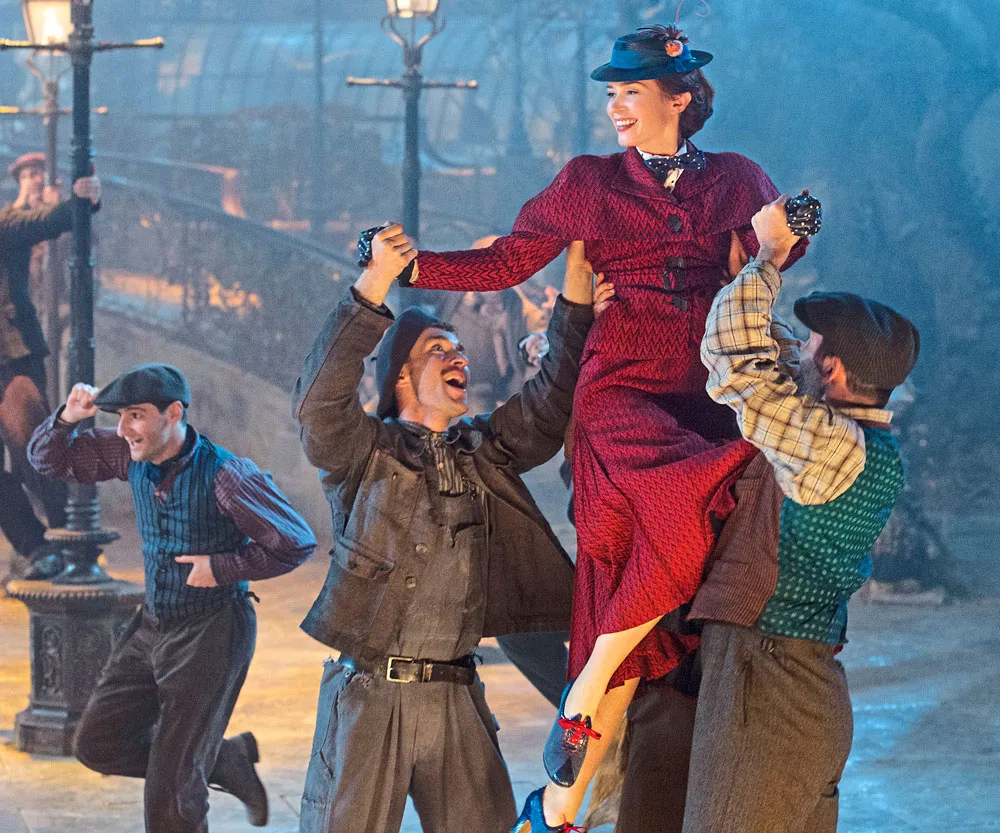 Mary Poppins in a red dress is joyfully lifted by dancers in a lively street scene with vintage attire and lampposts.