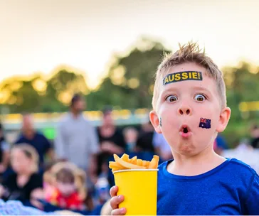 Child with 'Aussie' sticker and face paint holding fries, looking surprised at an outdoor event.