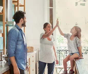 Family in a kitchen: a woman and a young girl high-five while a man watches and smiles.
