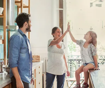 Family in a kitchen: a woman and a young girl high-five while a man watches and smiles.