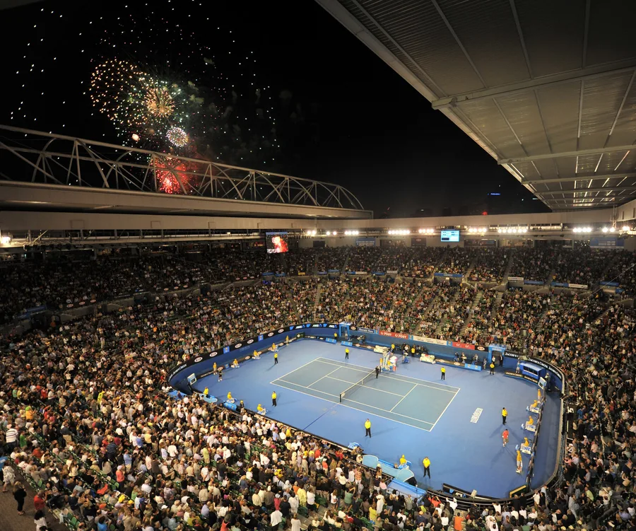A packed stadium in Melbourne during a night tennis match, with colorful fireworks lighting up the sky above.