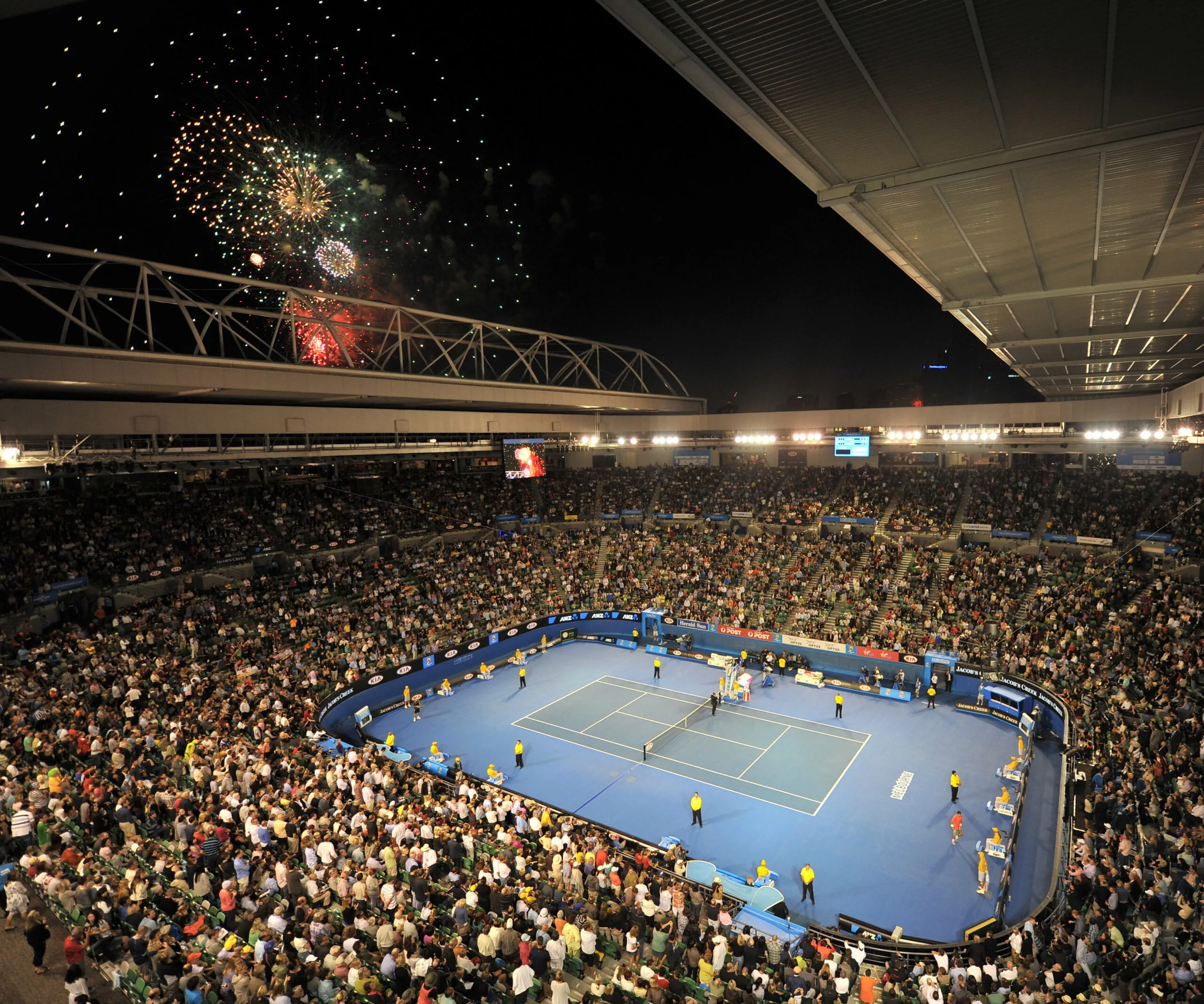 A packed stadium in Melbourne during a night tennis match, with colorful fireworks lighting up the sky above.