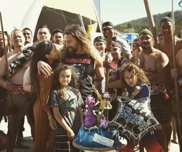 A group celebrates with a themed cake and balloons, surrounded by people in traditional attire outdoors.