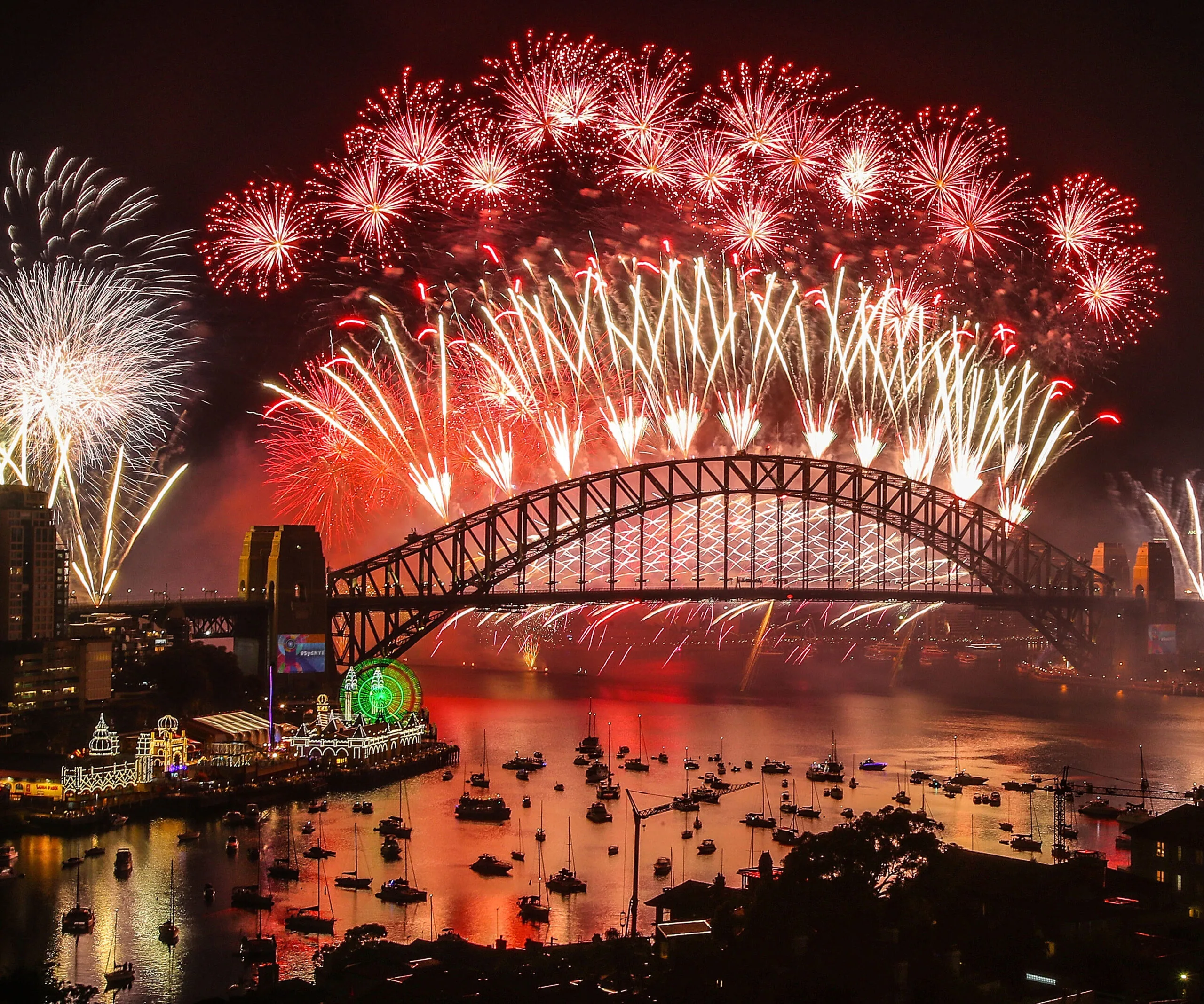 Fireworks light up the night sky over Sydney Harbour Bridge, reflecting in the water, with boats in the foreground.