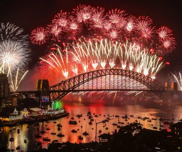 Fireworks light up the night sky over Sydney Harbour Bridge, reflecting in the water, with boats in the foreground.