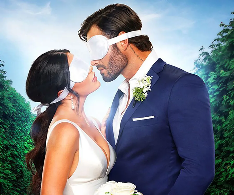 Bride and groom wearing blindfolds, about to kiss, in formal attire against lush green background.