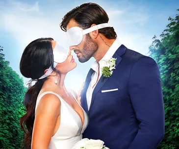 Bride and groom wearing blindfolds, about to kiss, in formal attire against lush green background.