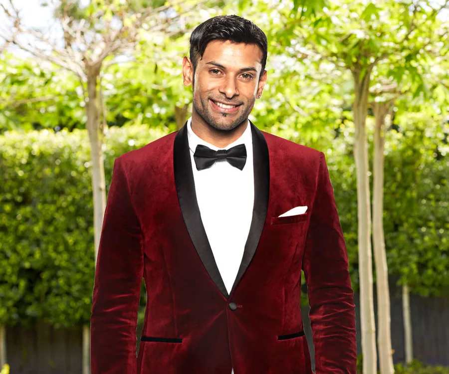 A man in a burgundy suit and bow tie stands smiling in a garden setting.