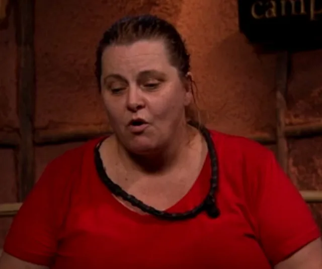 A person in a red shirt appears to be speaking or singing in a rustic setting.