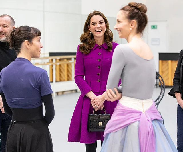 A woman in a purple coat talks to ballet dancers in a dance studio.