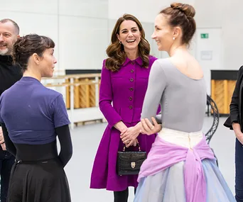 A woman in a purple coat talks to ballet dancers in a dance studio.