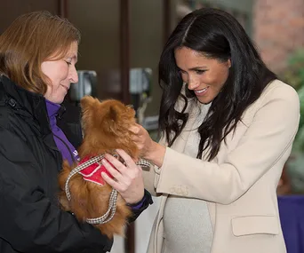 Woman in cream coat smiles while petting a small brown dog held by another woman in a black jacket.