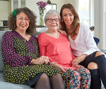Three women sitting together on a sofa, smiling warmly at the camera, in a cozy home setting.