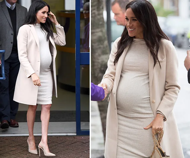 Pregnant woman in beige coat and dress, smiling while exiting a building and shaking hands outside.