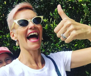 Person with short hair and sunglasses giving a thumbs-up outdoors, smiling energetically.