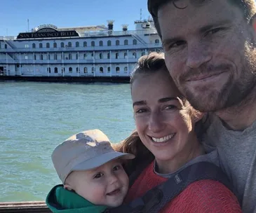 Family smiling by the water with a baby in a carrier; a large boat named "San Francisco Belle" is in the background.