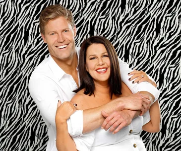 Two smiling people in white outfits hugging against a zebra print background.