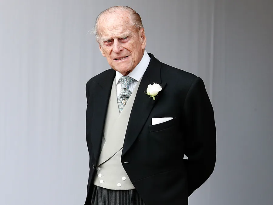 An elderly man in a formal suit with a white rose boutonniere stands in a light-colored setting.