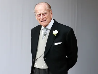 An elderly man in a formal suit with a white rose boutonniere stands in a light-colored setting.