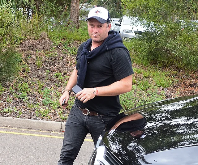 Man in casual attire with a cap, holding a phone beside a parked car, surrounded by greenery.