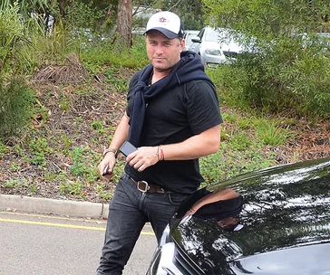 Man in casual attire with a cap, holding a phone beside a parked car, surrounded by greenery.