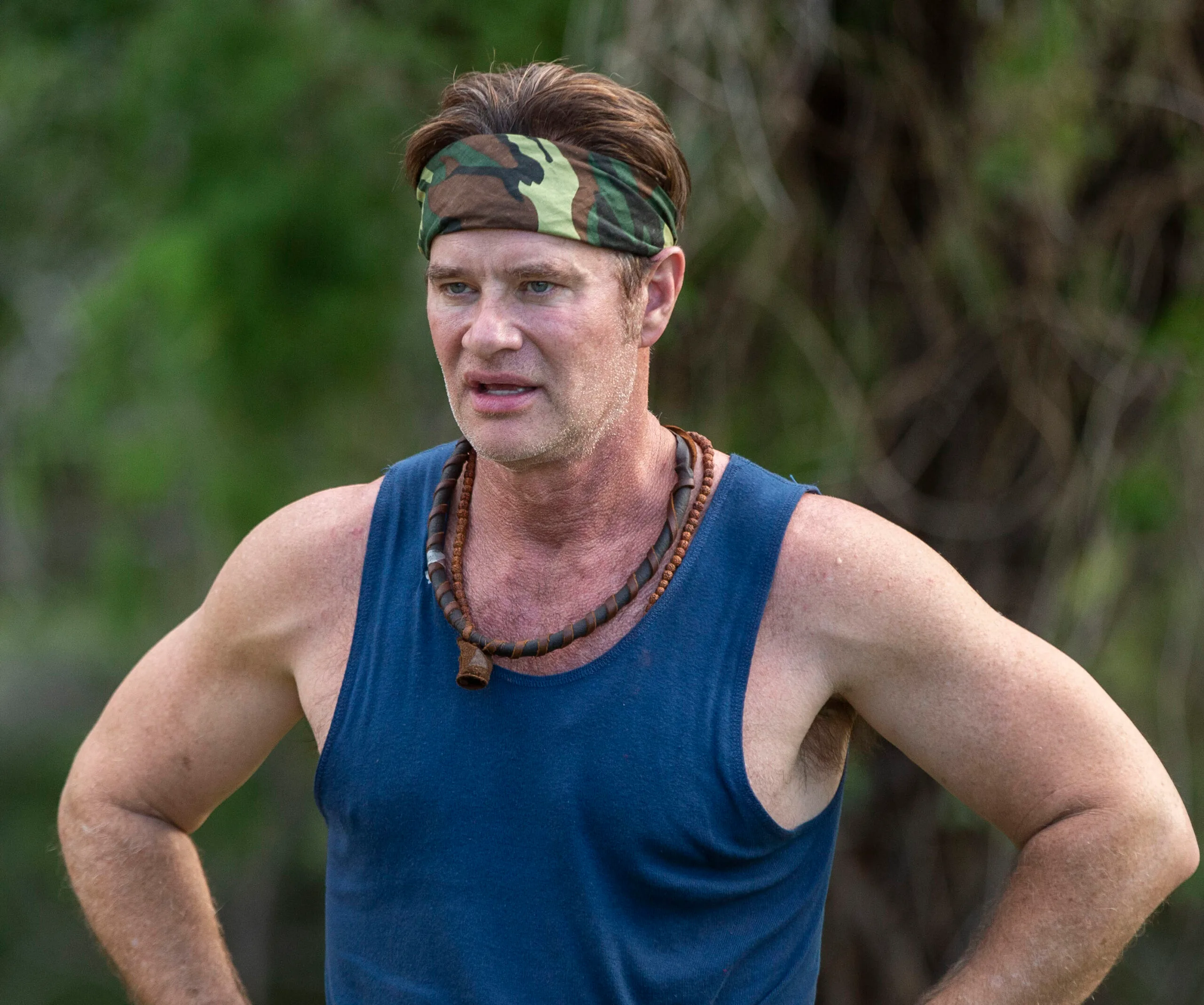 Man in a blue tank top and camouflage headband stands outdoors, arms on hips.