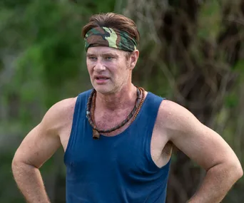 Man in a blue tank top and camouflage headband stands outdoors, arms on hips.