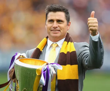 Man in suit with brown and yellow scarf holds a trophy, giving a thumbs up in a stadium setting.