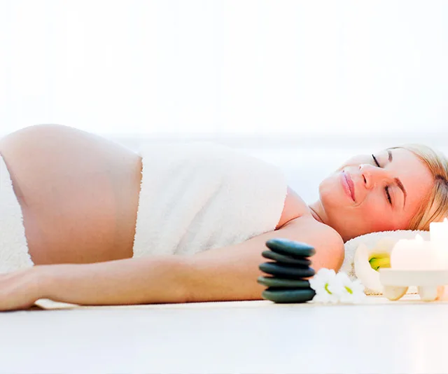 Pregnant woman relaxing at a spa, wrapped in a towel with stones, candles, and flowers nearby.