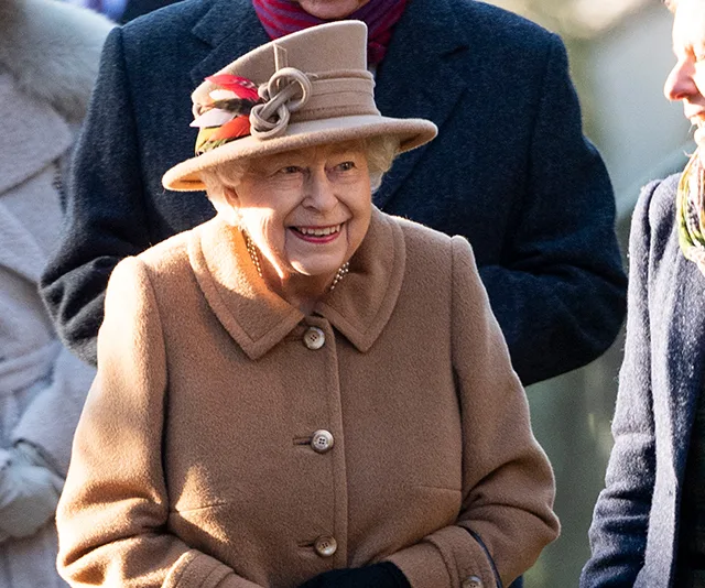 A smiling elderly woman in a tan coat and hat attends an outdoor event with others on a sunny day.