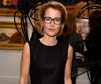 Gillian Anderson in a black dress and glasses at an indoor event, standing before artwork and floral decor.