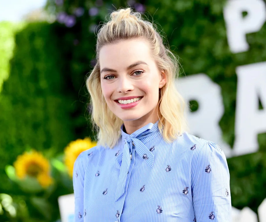 A woman with blonde hair in a blue patterned blouse smiles outdoors with greenery and sunflowers in the background.