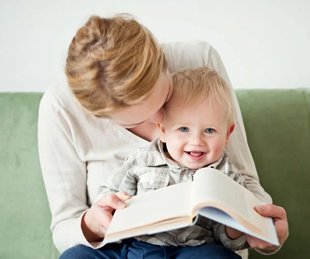 Mother reading a book to her smiling baby in her lap on a green sofa.