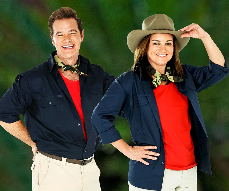 "Two people in jungle attire, a man with a scarf and woman with a hat, smiling against a green background."