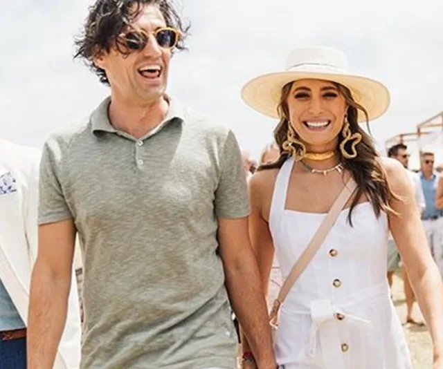 Couple smiling at an outdoor event; woman wearing a white dress and hat, and man in a green polo shirt with sunglasses.