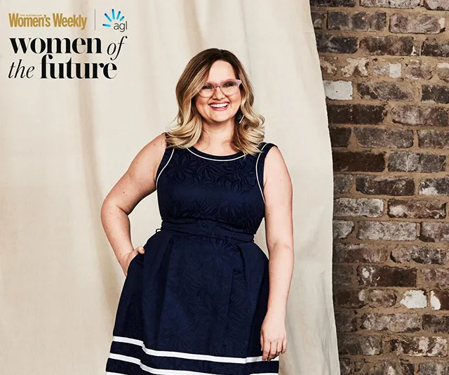 A smiling woman in a navy dress stands against a brick wall, featured in "Women of the Future" by Women's Weekly.