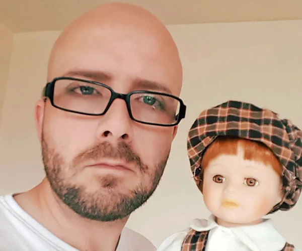 A man with glasses holding a doll wearing a plaid hat; both have serious expressions.