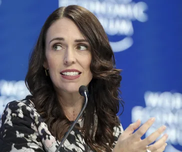 A person with long dark hair speaking at a conference, with a blurred blue background displaying "World Economic Forum."